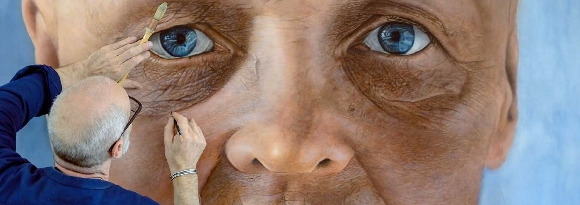 Auf dem Bild arbeitet der Maler Helmut Mair an einem riesigen, hyperrealistischen Porträt eines älteren Gesichts. Er selbst hat zwei Pinsel in den Händen und fügt sorgfältig Details in der Augenpartie des Gemäldes hinzu. Das Porträt zeigt ein stark vergrößertes Gesicht mit blauen Augen und vielen feinen Hautfalten.