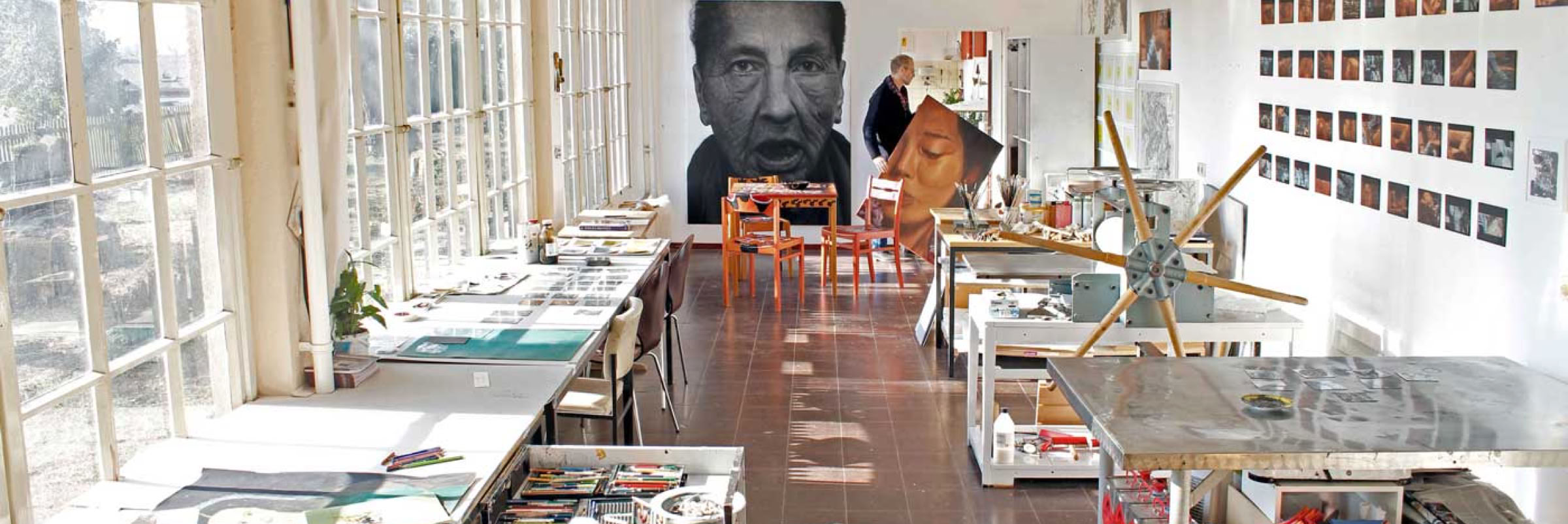 Ein heller Raum mit großen Fenstern auf der linken Seite, Tischen mit Kunstmaterialien, einem großen Porträt an der Wand und mehreren kleinen Bildern an der rechten Wand.